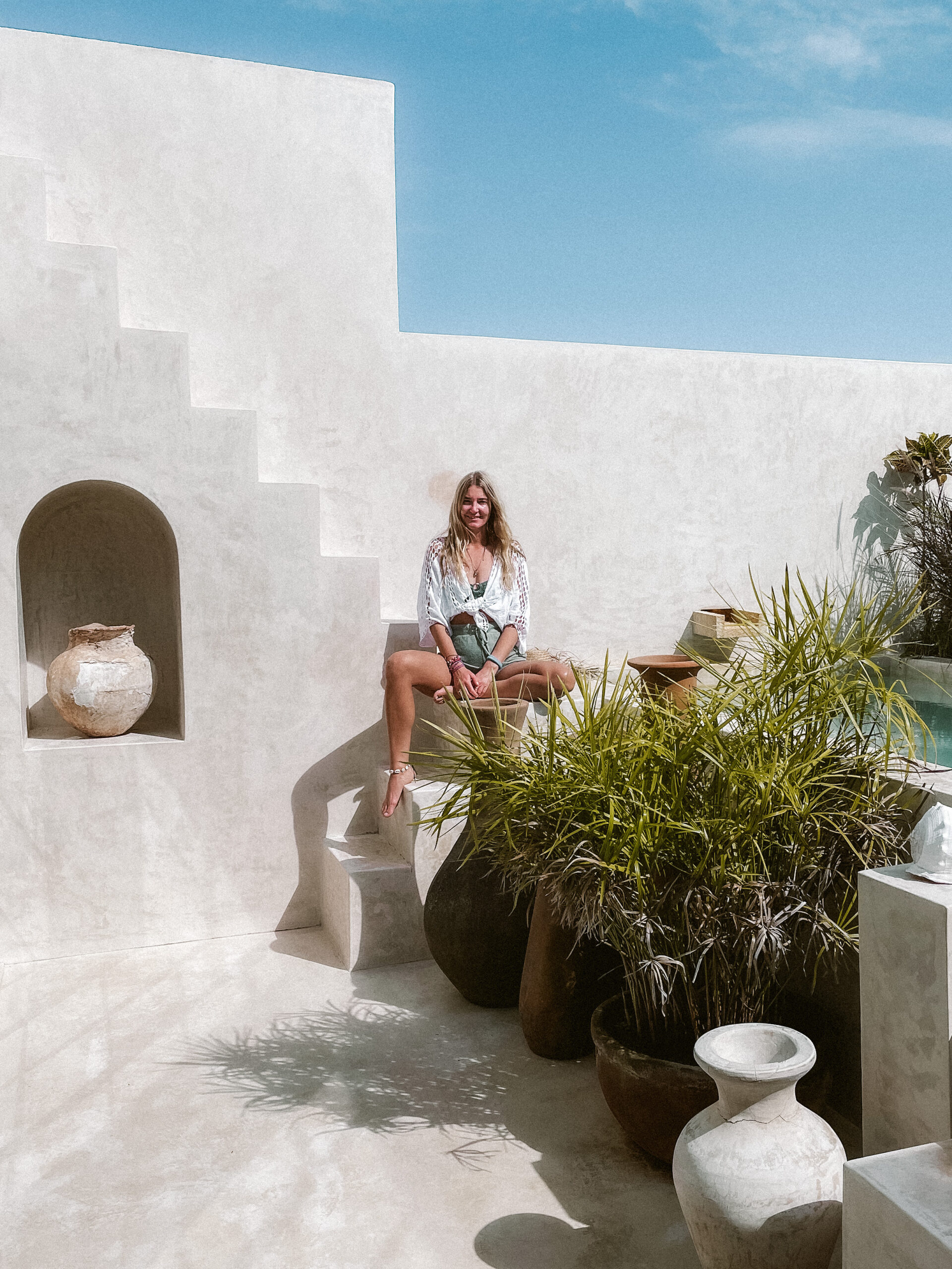 women sits in front of white wall with plants and pots in the background